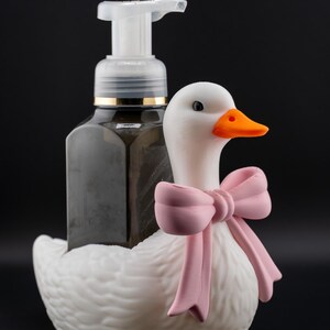 May include: A decorative soap dispenser featuring a white goose with a pink bow. The dispenser holds a dark liquid and has a clear pump. The goose has an orange beak and black eyes. The background is black.