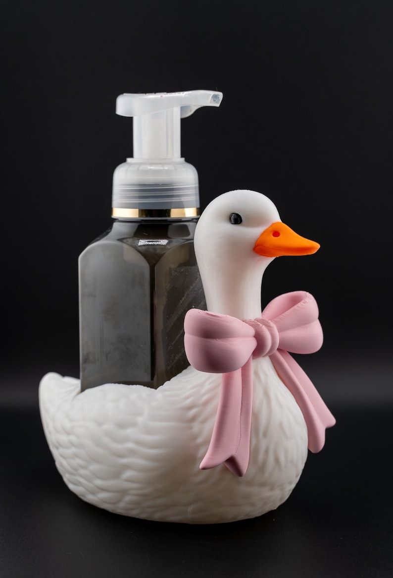May include: A decorative soap dispenser featuring a white goose with a pink bow. The dispenser holds a dark liquid and has a clear pump. The goose has an orange beak and black eyes. The background is black.