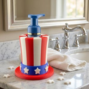 May include: A red, white, and blue soap dispenser shaped like a patriotic Uncle Sam hat. The dispenser has a blue pump and a red base with white star accents. The design is reminiscent of the American flag.