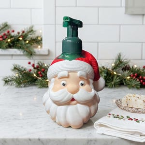 Könnte beinhalten: Ein Seifenspender in Form eines Weihnachtsmanns mit grünem Pumpkopf, rot-weißer Mütze und weißem Bart. Der Spender besteht aus geformtem Kunststoff und zeigt ein detailliertes Gesicht mit Augen, Nase und Schnurrbart. Der Pumpmechanismus ist dunkelgrün.