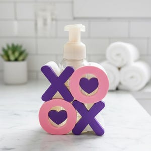 K&ouml;nnte beinhalten: Ein klarer Seifenspender mit wei&szlig;er Pumpe ist mit rosa und lila Holzbuchstaben verziert, die "XO" mit Herz-Ausschnitten buchstabieren. Der Spender steht auf einer wei&szlig;en Arbeitsplatte in einem Badezimmer.