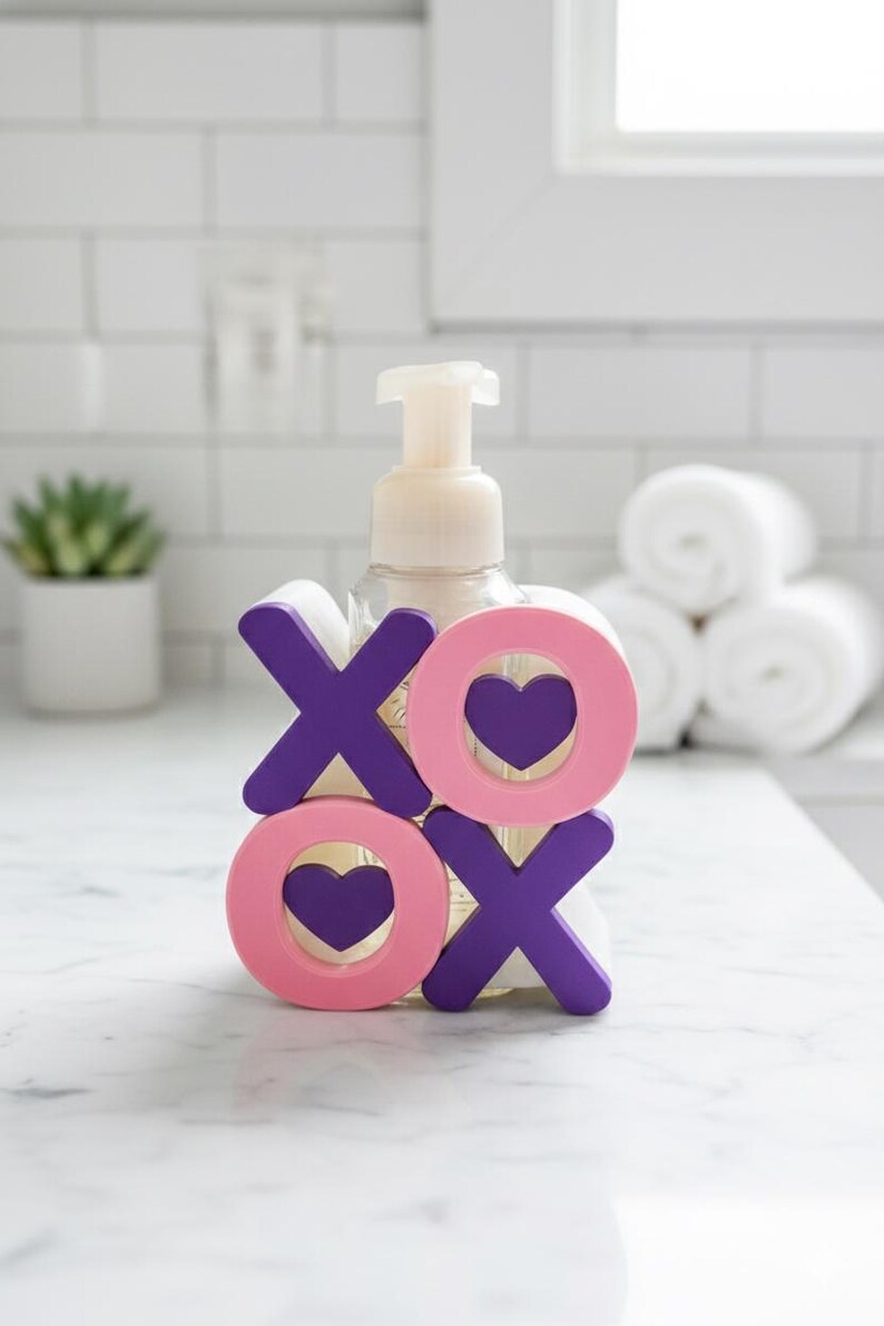 K&ouml;nnte beinhalten: Ein klarer Seifenspender mit wei&szlig;er Pumpe ist mit rosa und lila Holzbuchstaben verziert, die "XO" mit Herz-Ausschnitten buchstabieren. Der Spender steht auf einer wei&szlig;en Arbeitsplatte in einem Badezimmer.
