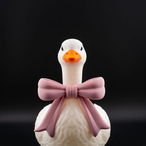May include: A white goose figurine with an orange beak and a pink bow. The goose has a textured body and is set against a black background. The bow is tied around the goose's neck.