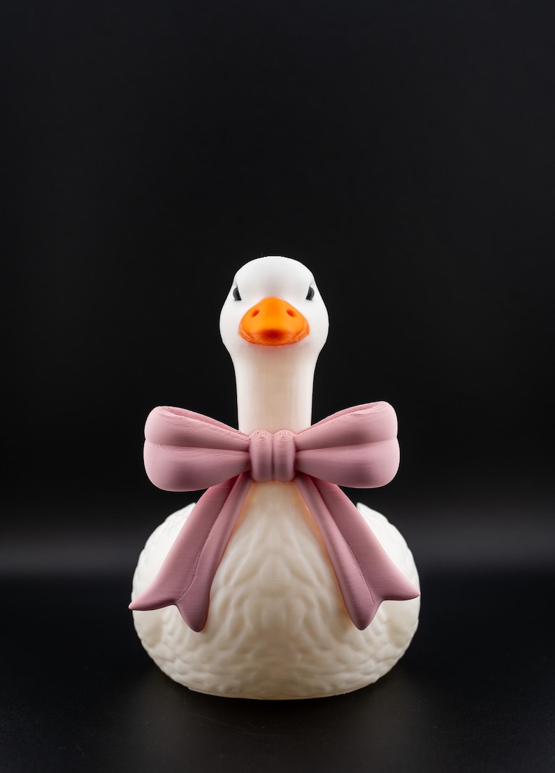 May include: A white goose figurine with an orange beak and a pink bow. The goose has a textured body and is set against a black background. The bow is tied around the goose's neck.