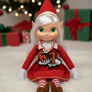Könnte beinhalten: Eine Weihnachtselfenpuppe mit weißem Haar, grünen Augen und spitzen Ohren. Die Puppe trägt ein rotes Kleid mit der Nummer 67, rot-weiß gestreifte Socken und braune Schuhe. Der Elf trägt einen roten Hut mit einem goldenen Band.