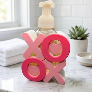 K&ouml;nnte beinhalten: Ein klarer Seifenspender mit beigefarbener Pumpe ist mit rosa und hellrosa XOXO-Buchstaben und Herzen verziert. Der Spender steht auf einer Marmorarbeitsplatte, im Hintergrund ein wei&szlig;es Handtuch und eine Sukkulente.
