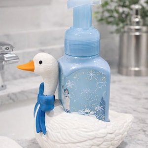 May include: A decorative soap dispenser featuring a white goose-shaped base with a blue bow. The light blue soap bottle has a winter scene design. The goose has an orange beak.