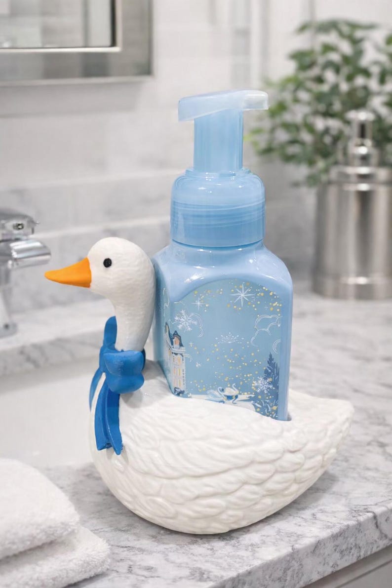 May include: A decorative soap dispenser featuring a white goose-shaped base with a blue bow. The light blue soap bottle has a winter scene design. The goose has an orange beak.