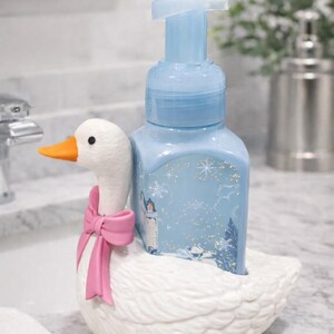 May include: A light blue soap dispenser with a winter scene design sits in a white ceramic goose-shaped holder. The goose has an orange beak and a pink bow. The dispenser is on a marble countertop.
