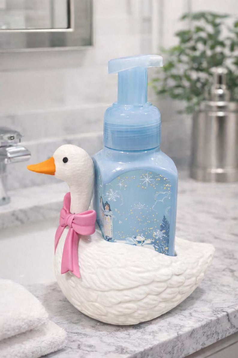 May include: A light blue soap dispenser with a winter scene design sits in a white ceramic goose-shaped holder. The goose has an orange beak and a pink bow. The dispenser is on a marble countertop.