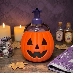 Könnte beinhalten: Ein Seifenspender in Form eines orangefarbenen Kürbisses mit einem schwarzen Jack-o'-Lantern-Gesicht. Der Spender verfügt über einen violetten Pumpmechanismus und einen schwarzen Griff. Der Kürbisbehälter besteht aus strukturiertem Material.