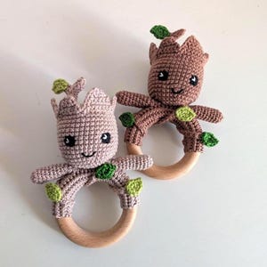 Könnte beinhalten: Zwei gehäkelte Babyrasseln, jede mit einem Groot-ähnlichen Charakter mit lächelndem Gesicht, kleinen Armen und grünen Blättern. Die Charaktere sind an einem Holzring befestigt. Einer ist hellbraun, der andere ist mauve.