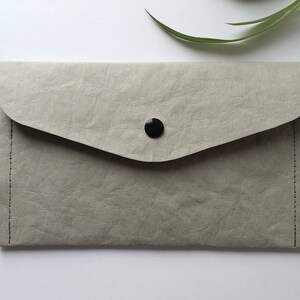 Puede incluir: Un bolso de mano gris claro con cierre de solapa y bot&oacute;n negro. El bolso tiene una superficie texturizada y una peque&ntilde;a leng&uuml;eta lateral. Los bordes est&aacute;n cosidos con hilo negro. Dise&ntilde;o sencillo y minimalista.