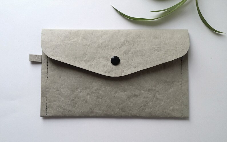 Puede incluir: Un bolso de mano gris claro con cierre de solapa y bot&oacute;n negro. El bolso tiene una superficie texturizada y una peque&ntilde;a leng&uuml;eta lateral. Los bordes est&aacute;n cosidos con hilo negro. Dise&ntilde;o sencillo y minimalista.