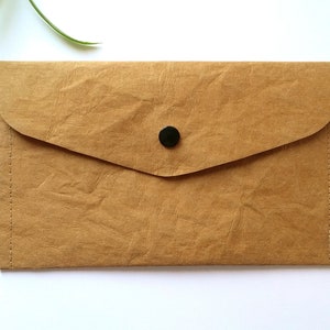 Puede incluir: Un clutch de papel marr&oacute;n con cierre de solapa y bot&oacute;n negro. El clutch tiene una peque&ntilde;a leng&uuml;eta lateral y est&aacute; cosido en los bordes. El fondo es blanco.
