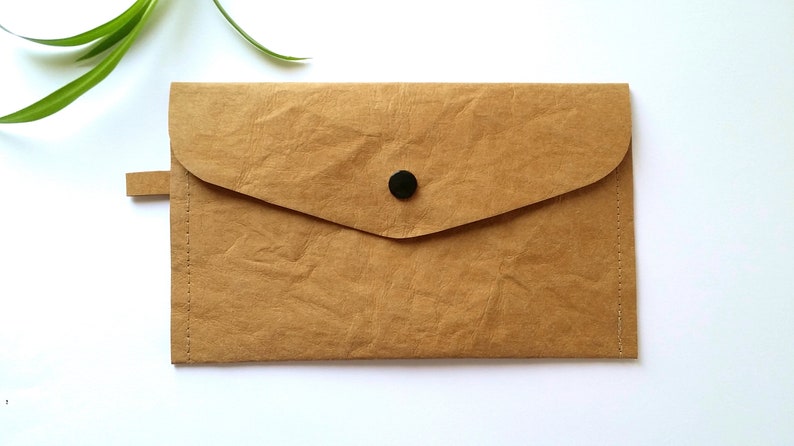 Puede incluir: Un clutch de papel marr&oacute;n con cierre de solapa y bot&oacute;n negro. El clutch tiene una peque&ntilde;a leng&uuml;eta lateral y est&aacute; cosido en los bordes. El fondo es blanco.