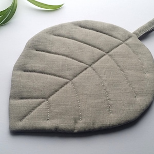 Puede incluir: Un posavasos de hoja gris con un asa de bucle. El posavasos est&aacute; hecho de una tela acolchada con una textura sutil.