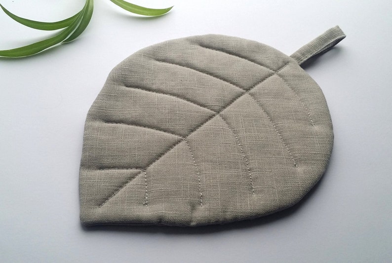 Puede incluir: Un posavasos de hoja gris con un asa de bucle. El posavasos est&aacute; hecho de una tela acolchada con una textura sutil.