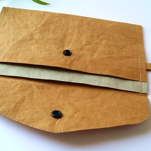 Puede incluir: Clutch de papel marr&oacute;n con cierre de solapa y dos cierres de bot&oacute;n a presi&oacute;n negros. El interior es de un tono beige m&aacute;s claro. El clutch tiene una peque&ntilde;a pesta&ntilde;a en el lateral y est&aacute; abierto, revelando el interior. El clutch est&aacute; sobre una superficie blanca.