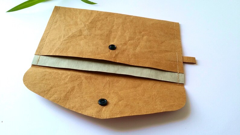 Puede incluir: Clutch de papel marr&oacute;n con cierre de solapa y dos cierres de bot&oacute;n a presi&oacute;n negros. El interior es de un tono beige m&aacute;s claro. El clutch tiene una peque&ntilde;a pesta&ntilde;a en el lateral y est&aacute; abierto, revelando el interior. El clutch est&aacute; sobre una superficie blanca.