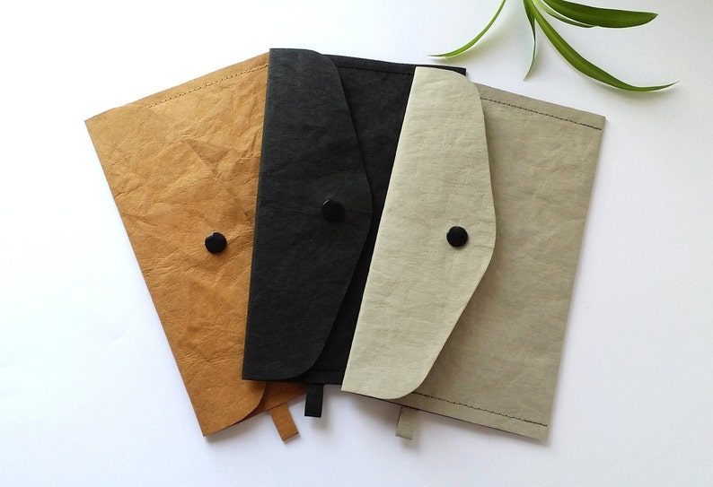 Puede incluir: Cuatro carteras de papel hechas a mano en tonos beige, negro y gris. Cada cartera tiene un cierre de solapa asegurado por un bot&oacute;n negro. Las carteras est&aacute;n dispuestas sobre una superficie blanca.