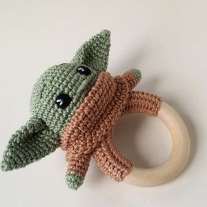 May include: A crocheted green and brown baby rattle with a wooden ring. The crocheted figure resembles a popular fictional character.