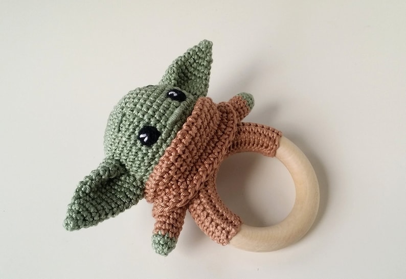 May include: A crocheted green and brown baby rattle with a wooden ring. The crocheted figure resembles a popular fictional character.