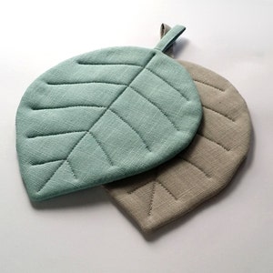 Puede incluir: Dos posavasos acolchados en forma de hoja, uno de color verde azulado claro y el otro de color beige claro.
