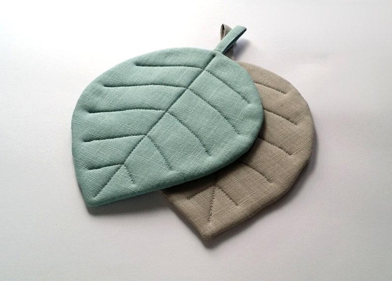 Puede incluir: Dos posavasos acolchados en forma de hoja, uno de color verde azulado claro y el otro de color beige claro.