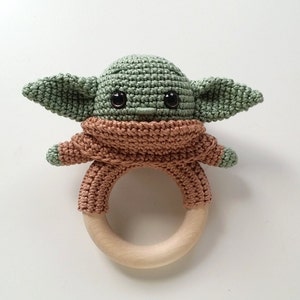 May include: A green crocheted baby rattle shaped like a character with large ears and big eyes. The rattle has a brown crocheted body and a wooden ring.