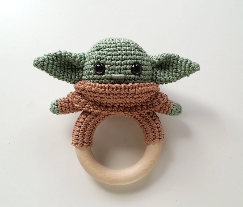 May include: A green crocheted baby rattle shaped like a character with large ears and big eyes. The rattle has a brown crocheted body and a wooden ring.