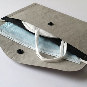 Puede incluir: Un estuche gris para mascarillas con interior negro, que contiene una mascarilla quir&uacute;rgica azul y blanca y una mascarilla blanca. El estuche tiene un cierre de bot&oacute;n a presi&oacute;n y un lazo. El estuche est&aacute; hecho de un material texturizado.
