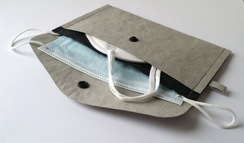 Puede incluir: Un estuche gris para mascarillas con interior negro, que contiene una mascarilla quir&uacute;rgica azul y blanca y una mascarilla blanca. El estuche tiene un cierre de bot&oacute;n a presi&oacute;n y un lazo. El estuche est&aacute; hecho de un material texturizado.