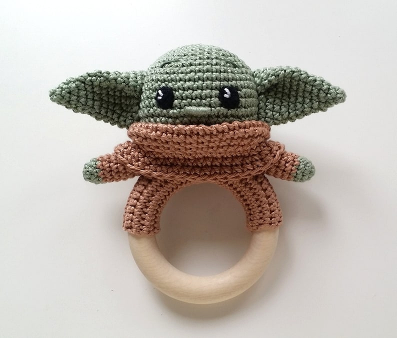 May include: A crocheted green and brown baby rattle shaped like a character from Star Wars. The rattle has a wooden ring and is a great toy for babies.