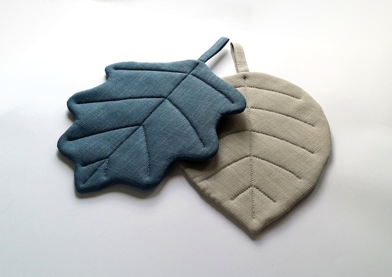 Puede incluir: Dos posavasos con forma de hoja, uno azul y otro beige, con un dise&ntilde;o acolchado. El posavasos azul tiene un asa de bucle.