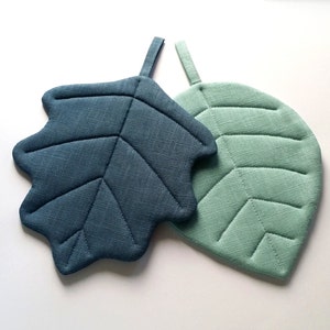 Puede incluir: Dos posavasos con forma de hoja, uno azul verdoso y otro verde claro, con textura acolchada.
