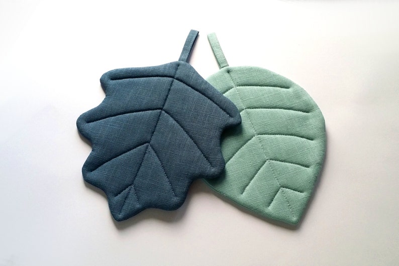 Puede incluir: Dos posavasos con forma de hoja, uno azul verdoso y otro verde claro, con textura acolchada.