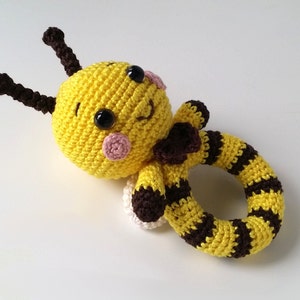 Puede incluir: Un sonajero para bebé en forma de abeja tejido a crochet con rayas amarillas y marrones. La abeja tiene una cara sonriente con ojos negros y mejillas rosadas.