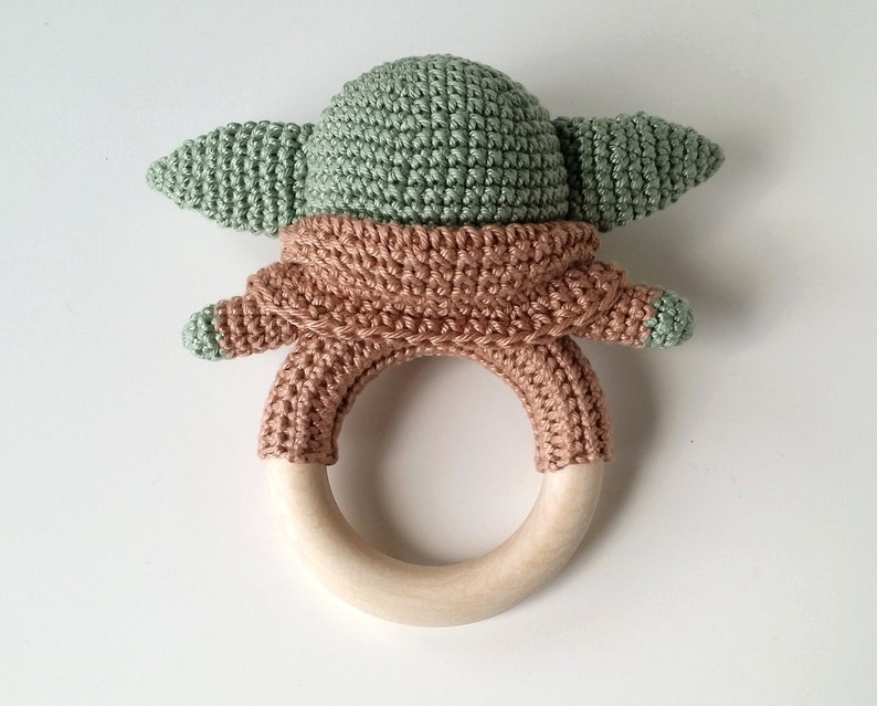 May include: A crocheted green and brown baby rattle with a wooden ring. The rattle is shaped like a character with large ears.