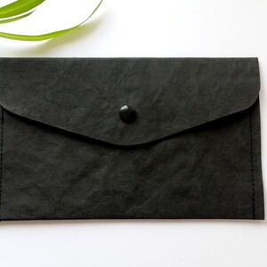Puede incluir: Un clutch negro estilo sobre con cierre de solapa y bot&oacute;n. El clutch est&aacute; hecho de un material texturizado y tiene una peque&ntilde;a pesta&ntilde;a en el lateral. Un tallo de planta verde est&aacute; en el fondo.