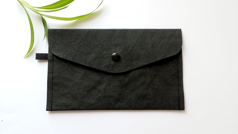 Puede incluir: Un clutch negro estilo sobre con cierre de solapa y bot&oacute;n. El clutch est&aacute; hecho de un material texturizado y tiene una peque&ntilde;a pesta&ntilde;a en el lateral. Un tallo de planta verde est&aacute; en el fondo.