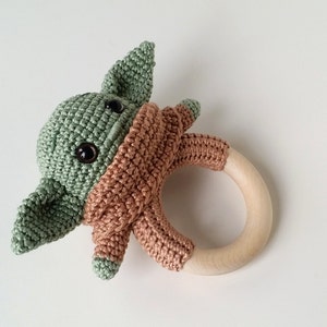 May include: A crocheted green and brown baby toy shaped like a character from the Star Wars franchise. The toy is attached to a wooden teething ring.