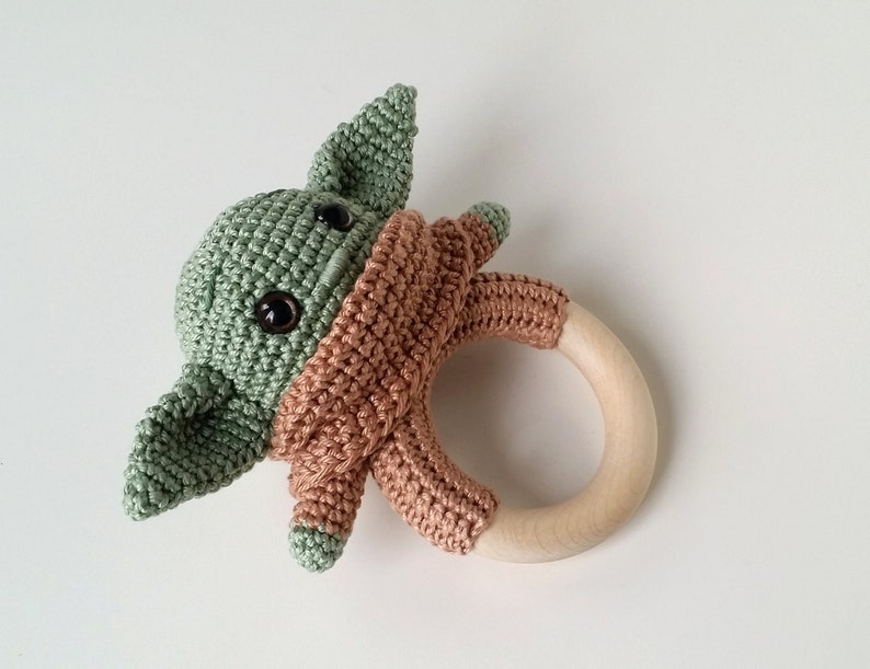 May include: A crocheted green and brown baby toy shaped like a character from the Star Wars franchise. The toy is attached to a wooden teething ring.