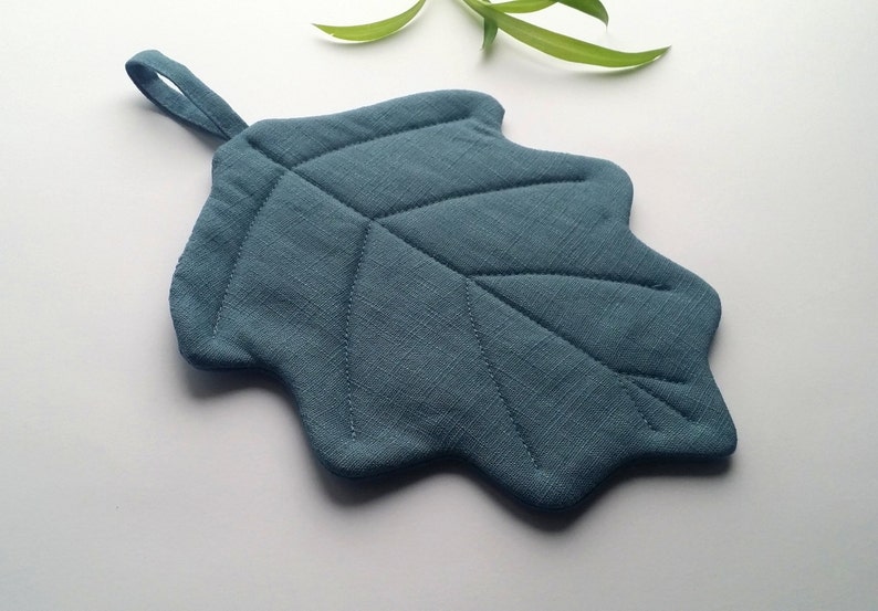 Puede incluir: Un posavasos en forma de hoja de color azul verdoso con un asa de bucle. El posavasos est&aacute; acolchado con un dise&ntilde;o de hoja.