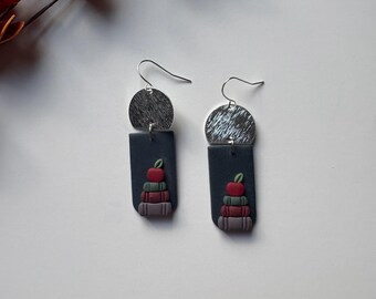 May include: A pair of handmade earrings featuring a silver-tone half-circle top and a dark gray rectangular base. The base has a stack of books with a red apple on top. The earrings are designed with a hook closure.