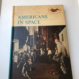 Puede incluir: Una cubierta de libro vintage con lomo verde y cubierta marrón. La cubierta presenta una foto de un astronauta con un traje espacial caminando por una calle. El título del libro es "Americans in Space" y el logotipo de la American Heritage Junior Library está en la esquina superior derecha.