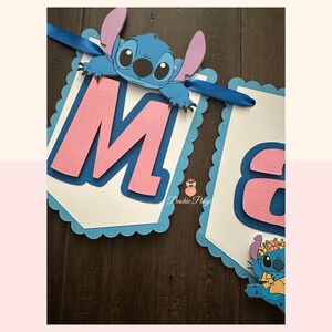 Puede incluir: Un cartel con la letra "M" en rosa, superpuesta sobre un fondo azul y un borde festoneado azul. Un personaje de dibujos animados azul est&aacute; adjunto en la parte superior. El cartel est&aacute; ensartado con una cinta azul. El segundo cartel tiene la letra "a".