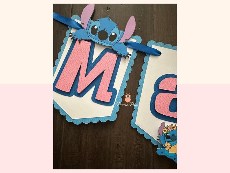 Puede incluir: Un cartel con la letra "M" en rosa, superpuesta sobre un fondo azul y un borde festoneado azul. Un personaje de dibujos animados azul est&aacute; adjunto en la parte superior. El cartel est&aacute; ensartado con una cinta azul. El segundo cartel tiene la letra "a".