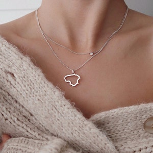 Puede incluir: Un collar de plata con dos cadenas finas. La cadena inferior presenta un colgante con la forma del contorno de un país. La cadena superior tiene una pequeña gema redonda. El collar se lleva sobre un suéter beige.