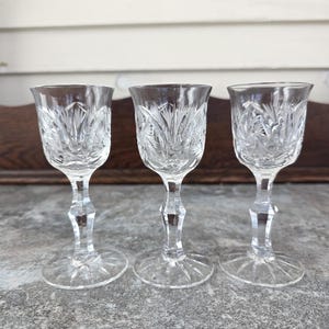 May include: Three clear crystal cordial glasses with intricate etched floral patterns. Each glass features a stemmed base and a detailed design on the bowl. The glasses are arranged in a row, showcasing their elegant craftsmanship.